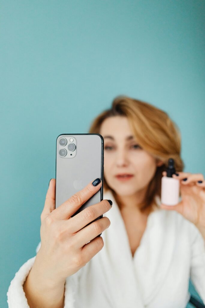 Adult woman in white gown taking a selfie while holding a cosmetic bottle.